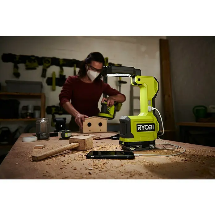Ryobi ONE+ LED-Arbeitsleuchte RMTL18-0 - 1.500 lm, Ohne Akku - Ein Ryobi-Arbeitsstrahler beleuchtet eine Werkbank mit einem Handy, einem Hammer und einer Person, die an einem Vogelhaus arbeitet.