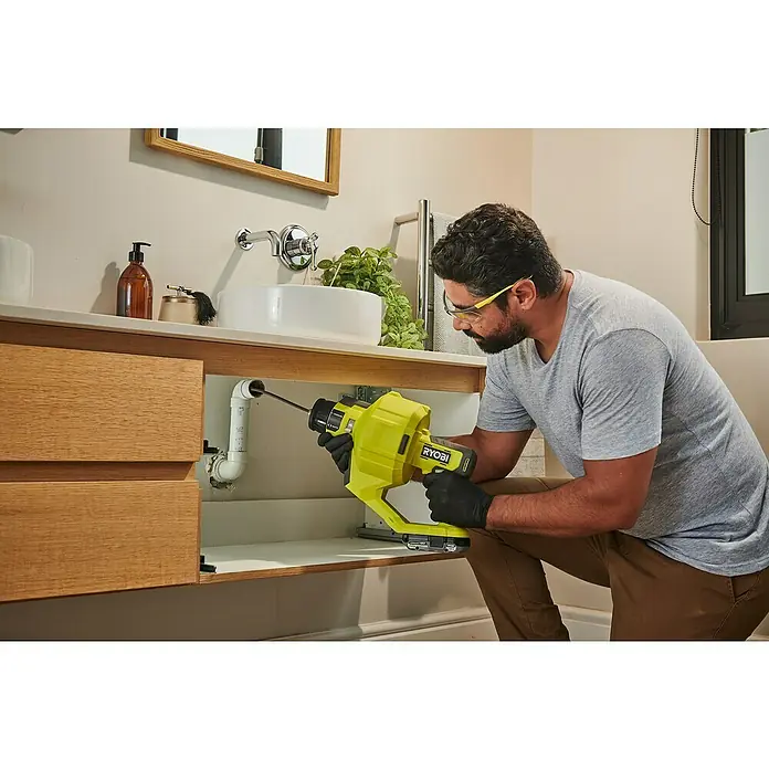 Ryobi ONE+ Akku-Rohrreinigungsgerät RDA18-0 - Ohne Akku - Ein Mann benutzt eine gelbe RYOBI Abflussreinigungsmaschine unter einem Badezimmerwaschbecken, um ein verstopftes Rohr zu reinigen.