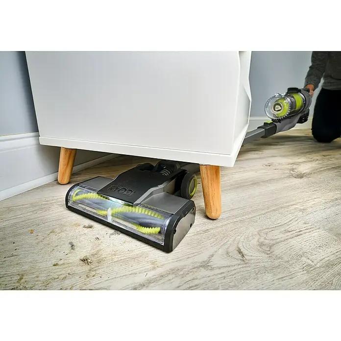 Ryobi ONE+ Akku-Handstaubsauger RSVS18-0 - 18 V, Ohne Akku - Nahaufnahme eines RYOBI-Staubsaugers, der unter einem weißen Nachttisch auf einem Holzboden saugt.