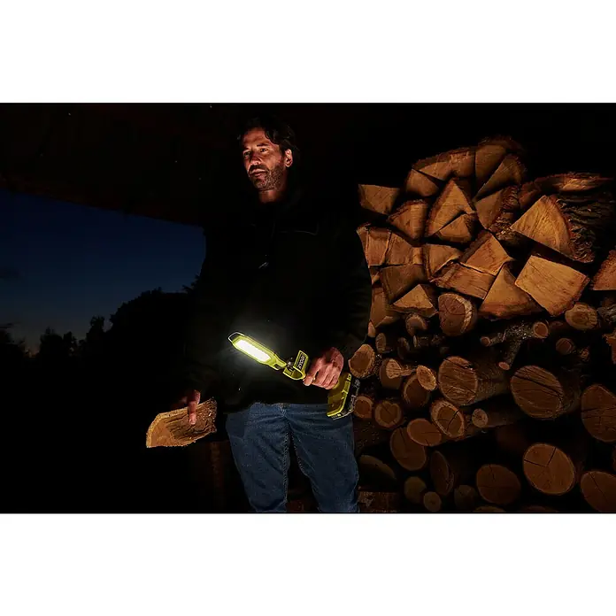 Ryobi ONE+ LED-Arbeitsleuchte SL18-0 - 18 V, Ohne Akku, 800 lm - Ein Mann hält eine leuchtende Ryobi-Arbeitsleuchte, die Holz neben einem Holzstapel beleuchtet, ideal für Arbeiten im Dunkeln.