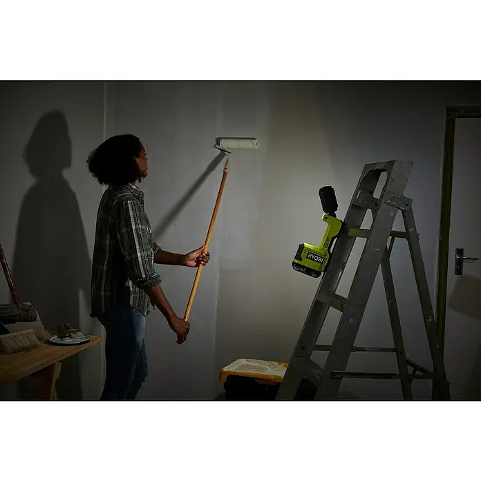 Ryobi ONE+ LED-Arbeitsleuchte RMTL18-0 - 1.500 lm, Ohne Akku - Eine Person streicht eine Wand mit einem Farbroller, während eine RYOBI Arbeitsleuchte an einer Leiter befestigt ist und Licht spendet.