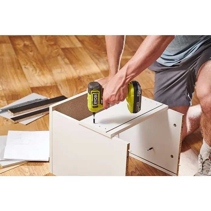 Ryobi ONE+ Akkuschrauber RSD18-0 - 18 V, Ohne Akku, 18 Nm - Ein Mann montiert ein weißes Möbelstück mit einem gelben Ryobi Akku-Bohrschrauber auf einem Holzboden. Werkzeuge liegen daneben bereit.