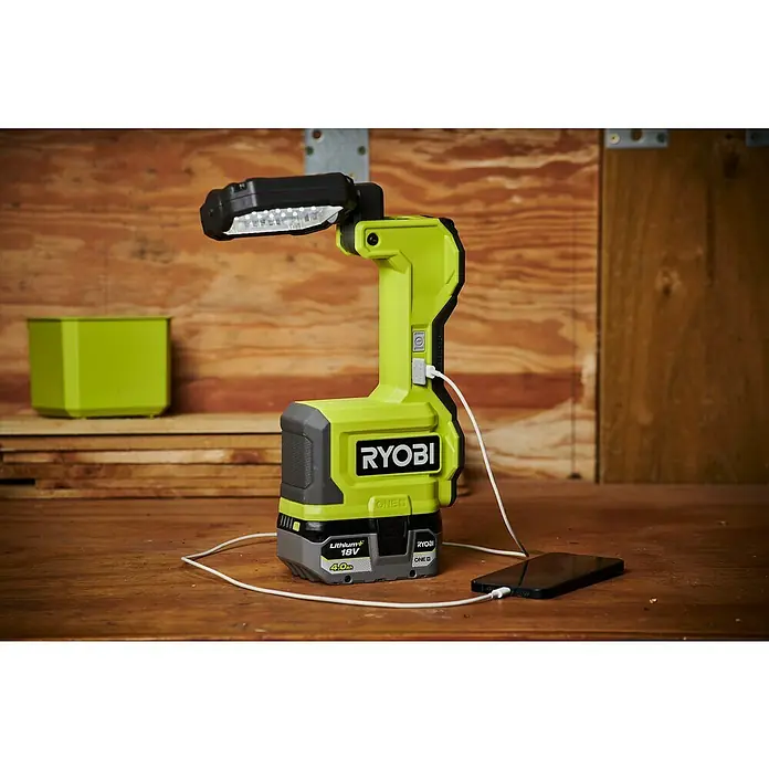Ryobi ONE+ LED-Arbeitsleuchte RMTL18-0 - 1.500 lm, Ohne Akku - Eine Ryobi Arbeitsleuchte mit Akku, die ein Smartphone auflädt, steht auf einem Holztisch.