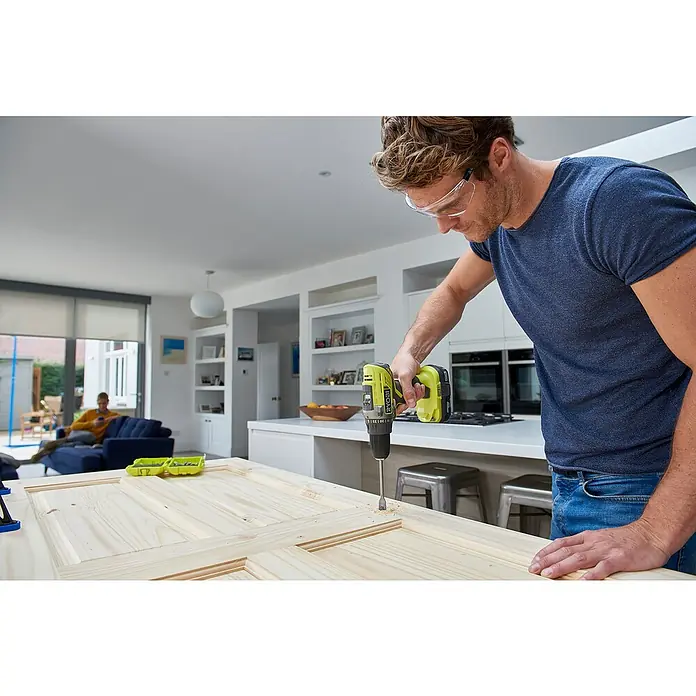 Ryobi ONE+ Maschinen-Set RCK184-252S - 18 V, 2 Akkus, 2 Ah - 5 Ah, 8 -tlg. - Ein Mann bohrt mit einem Ryobi Akku-Bohrschrauber Löcher in eine Holztür, während im Hintergrund eine Person auf einem Sofa sitzt.
