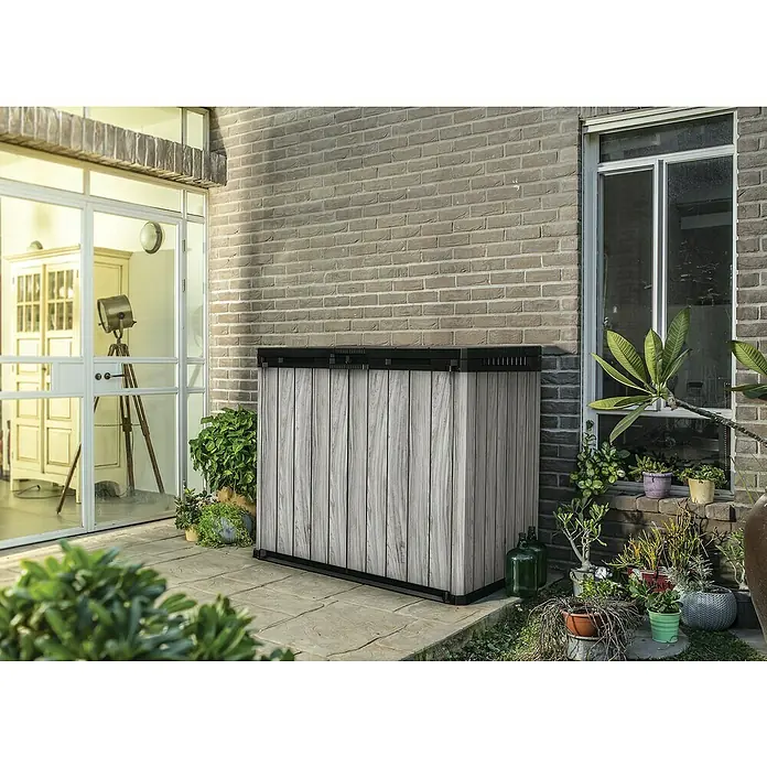 Keter Truhe Store it out Darwin - L x B x H: 80 x 140 x 118 cm, Stormgrey, Kunststoff - Außenaufnahme eines hölzernen Aufbewahrungsschranks mit schwarzem Rahmen neben einer Steinmauer und Pflanzen.