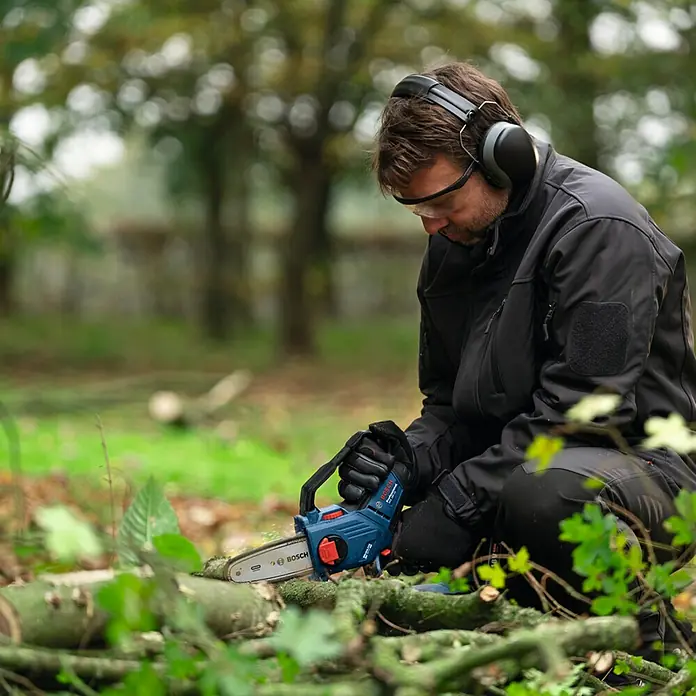 Bosch Professional AMPShare 18V Sägekette - Passend für: Bosch Professional AMPShare 18V Akku-Astsäge GKE 18V-15 - Ein Mann kniet draußen und benutzt eine kleine Bosch-Kettensäge, um Holz zu schneiden, während er Kopfhörer und eine Schutzbrille trägt.