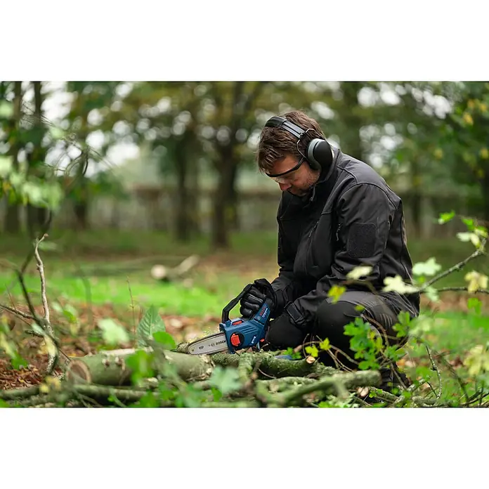 Bosch Professional AMPShare 18V Akku-Astsäge GKE 18V-15 - 18 V, Ohne Akku, Schwertlänge: 15 cm - Mann hockt im Freien und schneidet Holz mit einer kompakten Bosch Akku-Kettensäge, während er Schutzausrüstung trägt.