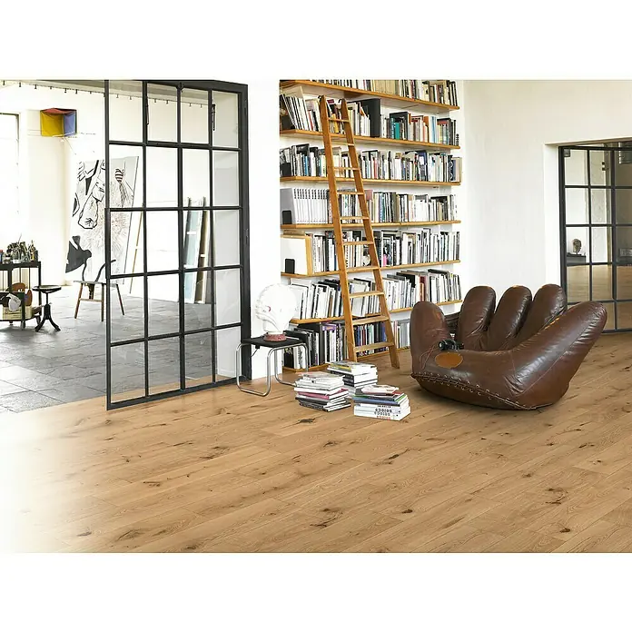 Parador Parkett Basic 11.5 - Eiche Chianti leicht geweißt, Landhausdiele, 1.080 x 185 x 11,5 mm - Heller Wohnraum mit Holzboden. Ein brauner Sessel in Form eines Baseballhandschuhs und ein Bücherregal mit einer Leiter sind zu sehen.