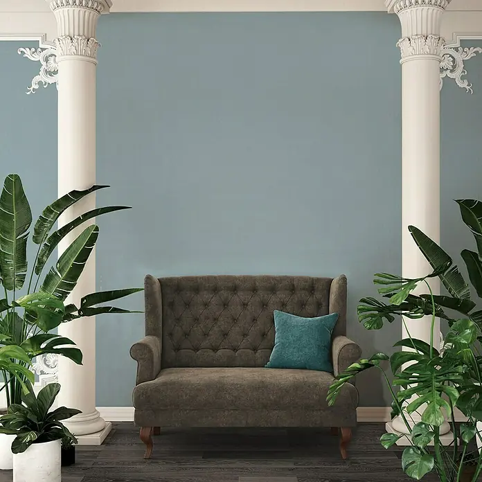 A.S. Création Ethnic Origin Vliestapete I - Blaugrau, Uni, 10,05 x 0,53 m - Elegantes Sofa mit braunem Bezug und blauem Kissen, platziert zwischen Säulen und üppigen Pflanzen, strahlt eine klassische und komfortable Atmosphäre aus.