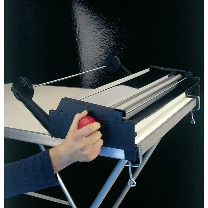 Decora Tapetenkleistergerät - Geeignet für: Euro Rollen 53 cm - Eine Person bedient eine Rollenschneidemaschine, um Papier präzise zuzuschneiden. Die Maschine steht auf einem Tisch mit silbernen Beinen.