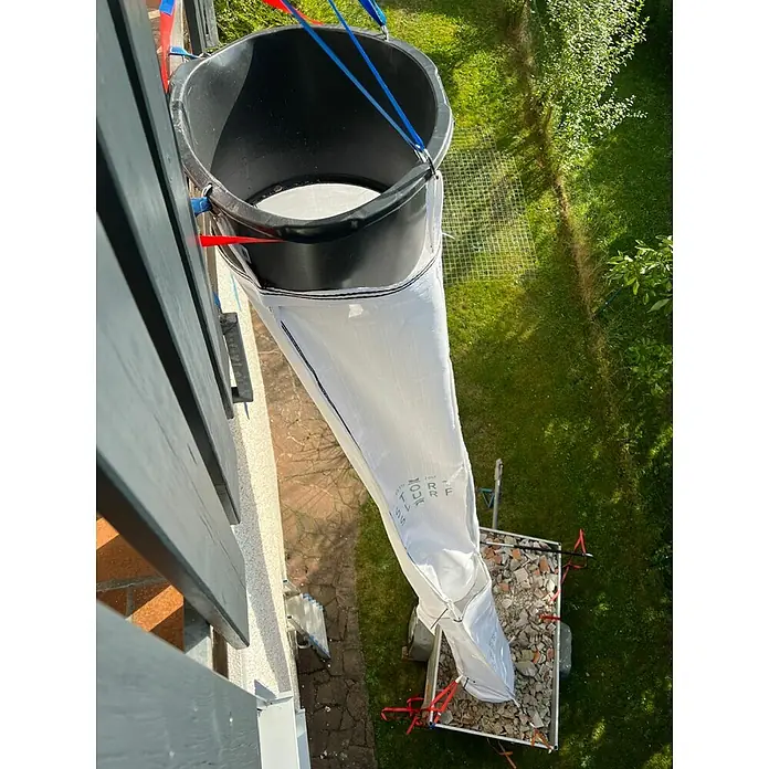 Stortslurf Trichter - Durchmesser: 50 cm - Eine Müllrutsche, bestehend aus einem Eimer und einem Sack, hängt an einem Balkon und führt zu einem Anhänger voller Bauschutt.