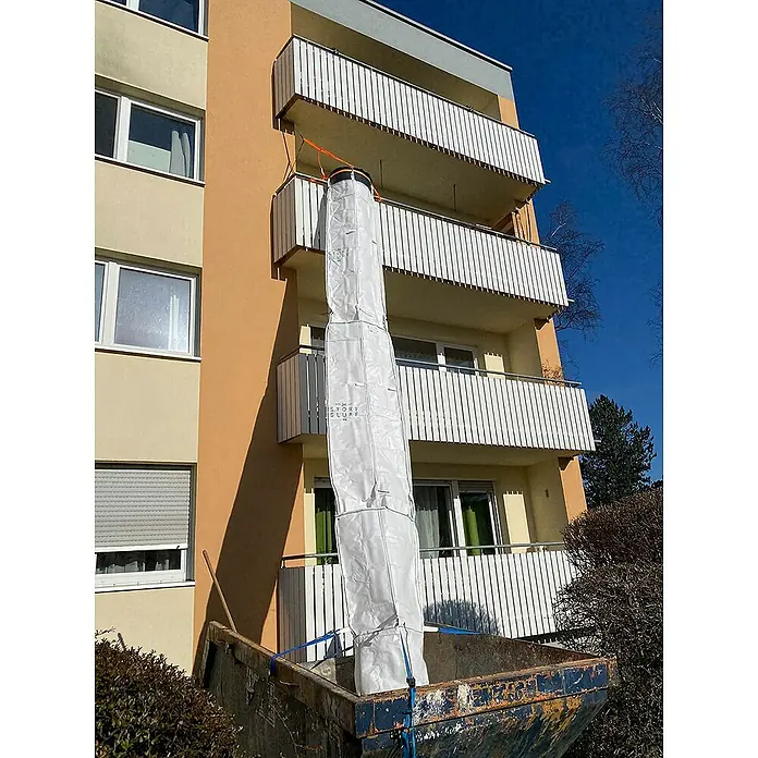 Stortslurf Schuttrutsche - Ø x L: 50 x 300 cm - Das Bild zeigt einen Bauschuttsack, der von einem Balkon eines Mehrfamilienhauses in einen Container abgeseilt wird. Der Sack ist weiß und mit Gurten gesichert.