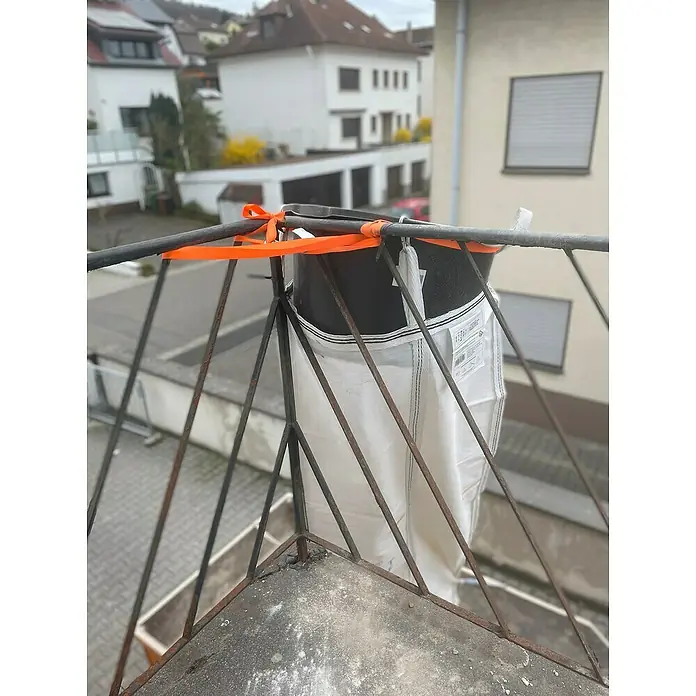 Stortslurf Trichter - Durchmesser: 50 cm - Ein großer Big Bag, gesichert mit einem orangefarbenen Gurtband, hängt an einem Balkongitter. Die Tasche ist schwarz-weiß und teilweise gefüllt.