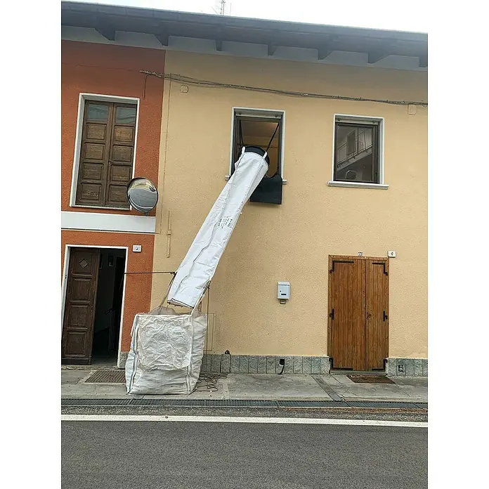 Stortslurf Schuttrutsche - Ø x L: 50 x 300 cm - Eine Abwurfvorrichtung für Bauschutt, die aus einem Fenster ragt und in einem großen, weißen Sack auf der Straße mündet.