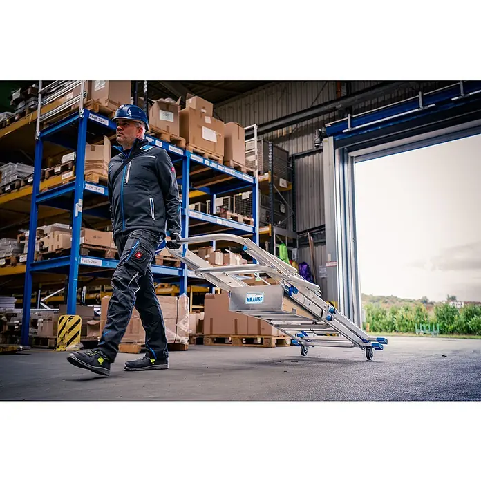 Krause Sprossen-Gelenk-Teleskopleiter Skymatic 5-9 Sprossen - Arbeitshöhe: 4,35 m, Anzahl Tritte: 9 Stk., Aluminium - Ein Bauarbeiter in Arbeitskleidung zieht einen Krause-Rollwagen mit einer Leiter in einem Lagerhaus.