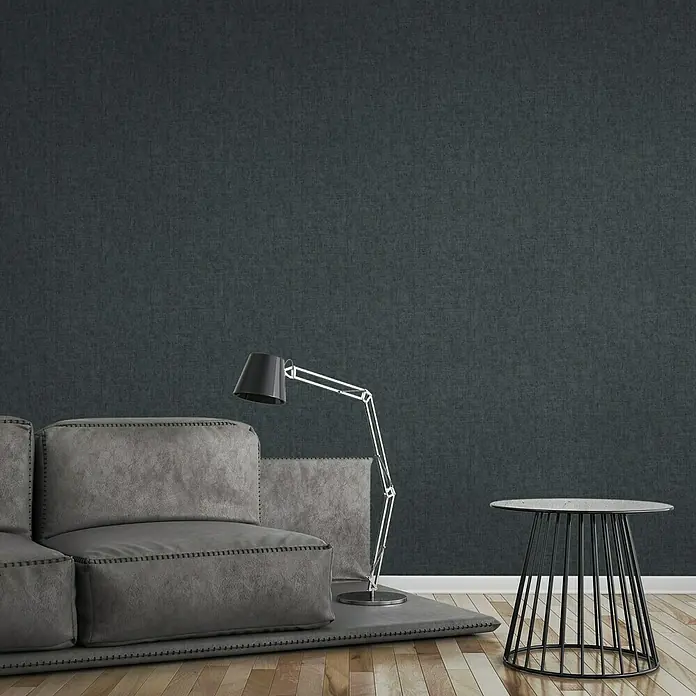 A.S. Création Greenery Vliestapete Strukturuni - Blau, Uni, 10,05 x 0,53 m - Ein gemütliches Wohnzimmer mit einem grauen Sofa, einer Stehlampe und einem Beistelltisch vor einer strukturierten, dunklen Wand.