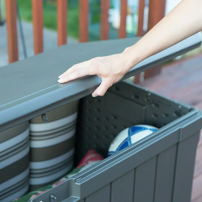 Lifetime Auflagenbox Harmony - 128 x 64 x 67 cm, Kunststoff, Braun - Eine Person öffnet eine graue Gartenbox gefüllt mit Kissen und einem Ball auf einer Holzterrasse.