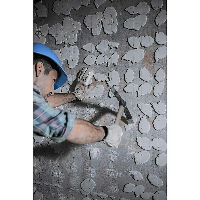 Bellota Martillo de albañil 26,5 cm - Largo: 40 mm, 500 g, Acero - Un trabajador de la construcción con casco azul usa un pico para quitar azulejos viejos de una pared, preparándola para otros nuevos.