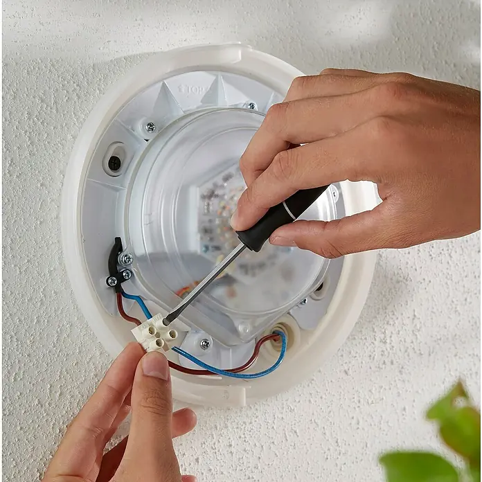 Philips Hue LED-Außenwandleuchte White & Color Ambiance Daylo - 15 W, 22 x 22 x 7,6 cm, Silber, IP44 - Eine Nahaufnahme einer Person, die eine LED-Leuchte mit einem Schraubenzieher an einer weißen Wand befestigt. Die Leuchte hat eine klare Abdeckung.