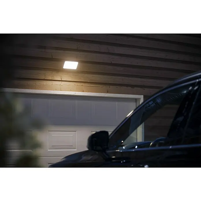 Philips Wiz LED-Außenwandleuchte Welcome - 20,5 W, 16 x 2 x 15,3 cm, Schwarz, IP44 - Ein beleuchteter, quadratischer Aussenleuchte beleuchtet eine Garage und ein davor geparktes Auto und sorgt für Sicherheit und Komfort im Aussenbereich.