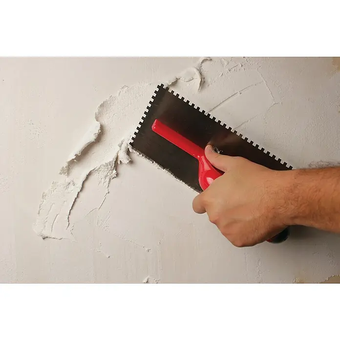 Rubi Llana dentada - 120 x 280 mm - Primer plano de una persona que aplica compuesto para paneles de yeso a una pared con una llana de mango rojo, creando una superficie texturizada.