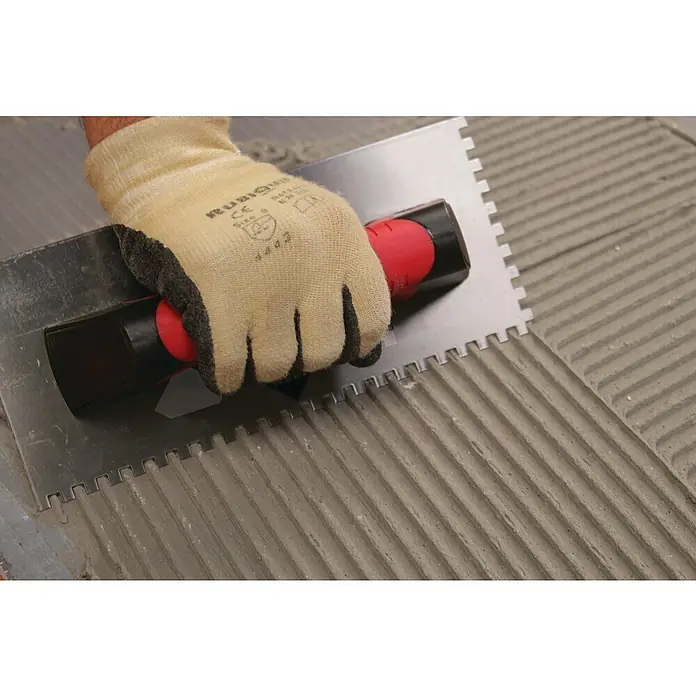 Rubi Llana dentada Rubiflex - 120 x 280 mm, Dientes 10x10, Mango bimaterial - Primer plano del trabajo de alicatado con una llana dentada, sujetada por una mano con un guante de trabajo.