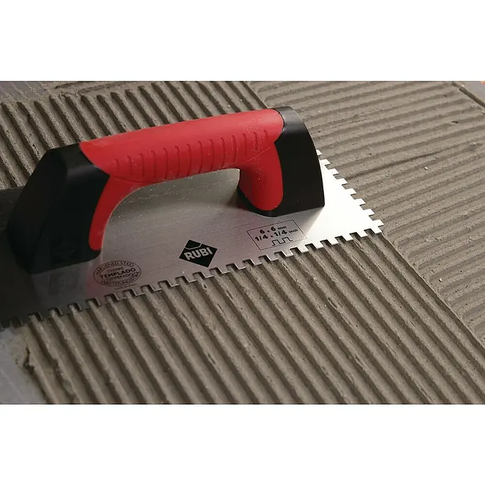 Rubi Llana dentada Rubiflex - 120 x 280 mm, Dientes 10x10, Mango bimaterial - Primer plano de una llana dentada RUBI con mango rojo, descansando sobre una superficie adhesiva acanalada para azulejos.