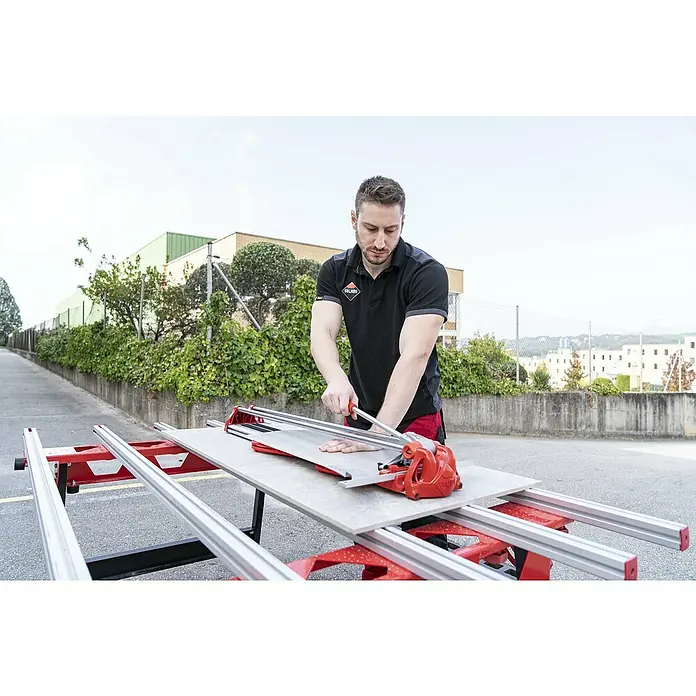 Rubi Cortador manual de azulejos Fast 65 - Longitud de corte máx.: 650 mm, Espesor máx. de azulejo: 12 mm - Un hombre corta azulejos con un cortador de azulejos RUBI. La herramienta es roja y plateada y está colocada sobre una mesa.
