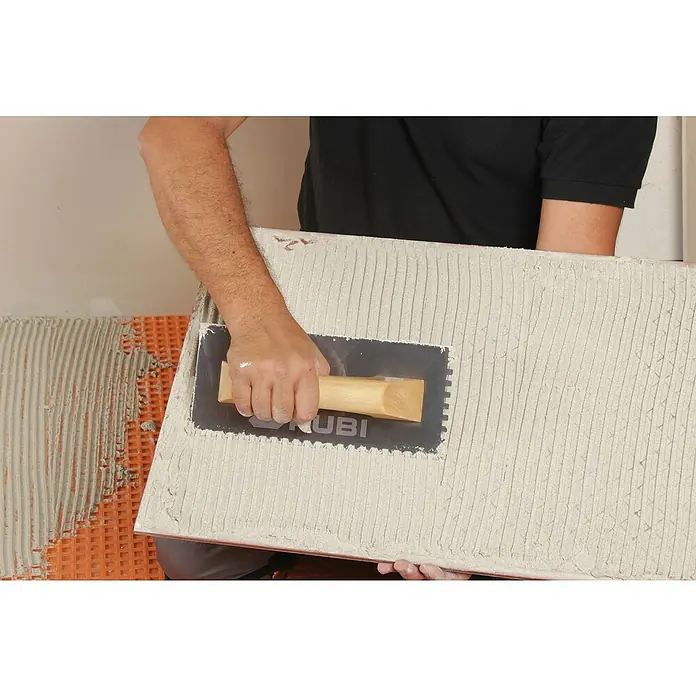 Rubi Llana dentada - 120 x 280 mm, Mango de madera, Dientes 6x6 - Un alicatador aplica adhesivo a un azulejo con una llana RUBI para su colocación en una pared ya preparada con adhesivo para azulejos.