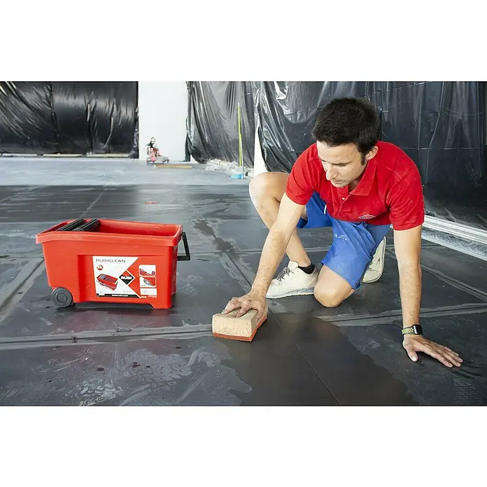 Rubi Esponja multiuso Mixta Superpro - L x An x Al: 20 x 13 x 7 cm - Un hombre arrodillado junto a un cubo rojo RubiClean está limpiando un piso de baldosas negras con una esponja.
