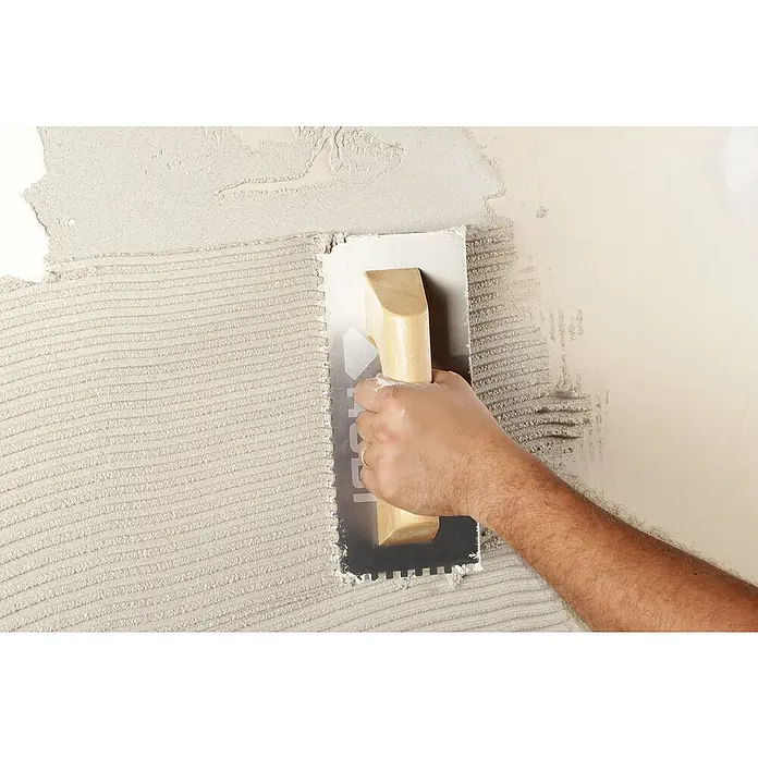 Rubi Llana dentada - 120 x 280 mm, Mango de madera, Dientes 6x6 - Primer plano de un manitas aplicando adhesivo para baldosas a una pared con una llana para colocar baldosas profesionalmente.