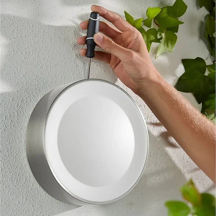 Philips Hue LED-Außenwandleuchte White & Color Ambiance Daylo - 15 W, 22 x 22 x 7,6 cm, Silber, IP44 - Nahaufnahme einer runden Deckenleuchte mit silbernem Rand, die mit einem Schraubendreher an einer Wand befestigt wird.