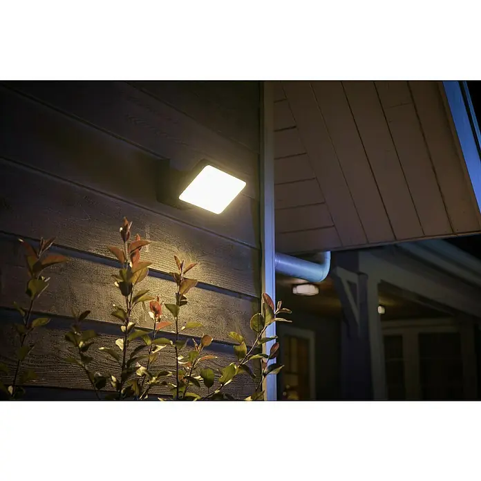 Philips Wiz LED-Außenwandleuchte Welcome - 20,5 W, 16 x 2 x 15,3 cm, Schwarz, IP44 - Eine quadratische Außenleuchte spendet helles Licht an der Hauswand und beleuchtet eine Pflanze im Vordergrund.