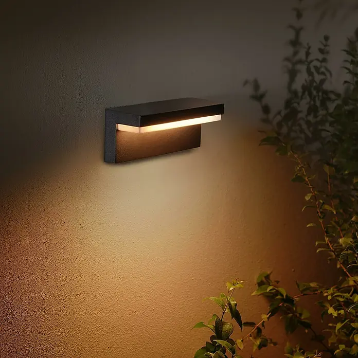 Philips Hue LED-Außenwandleuchte Nyro - 13,5 W, 25,4 x 88 x 10,1 cm, Schwarz, IP44 - Moderne schwarze Wandleuchte beleuchtet eine strukturierte Wand und wirft warmes Licht. Dekorative Aussenbeleuchtung mit Pflanzen im Hintergrund.