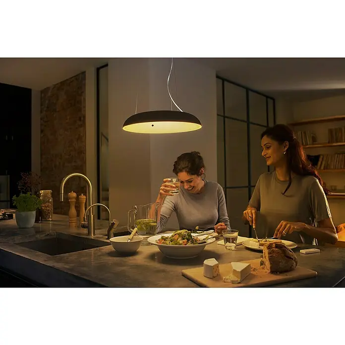 Philips Hue LED-Pendelleuchte rund White Ambience Amaze - 25 W, Ø x H: 43,4 x 150 cm, Schwarz - Diese Aufnahme zeigt eine moderne Philips Pendelleuchte über einem Esstisch, an dem sich zwei Personen unterhalten. Das Licht ist warm und einladend.