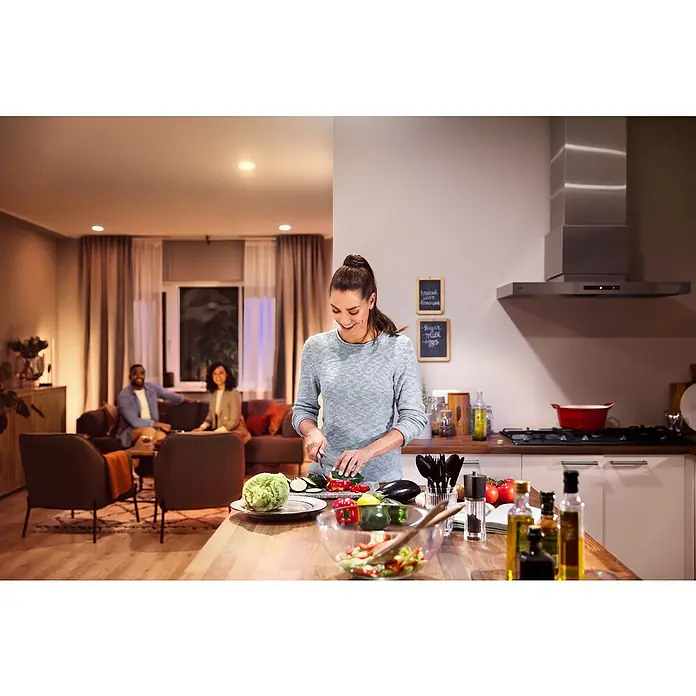 Philips Hue Smart-LED-Lampen Set White - E27, Dimmbar, Warmweiß, 806 lm, 9,5 W, 2 Stk. - Eine Frau bereitet mit einem Philips Produkt in einer Küche einen Salat zu, während sich im Hintergrund Personen im Wohnzimmer aufhalten.