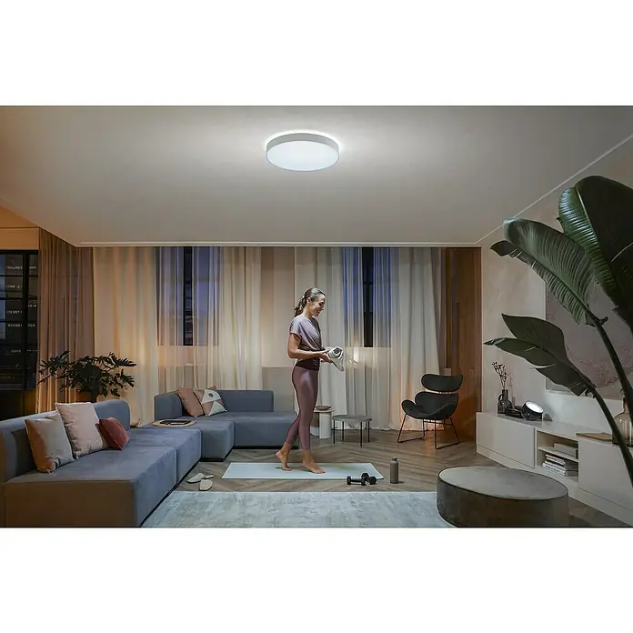 Philips Hue LED-Deckenleuchte rund White Ambience Enrave XL - 48 W, Ø x H: 55,1 x 8,4 cm, Weiß, Kaltweiß - Eine moderne Deckenleuchte erhellt einen Wohnraum mit einer Frau beim Sport, ein minimalistisches Design für stilvolle Beleuchtung.
