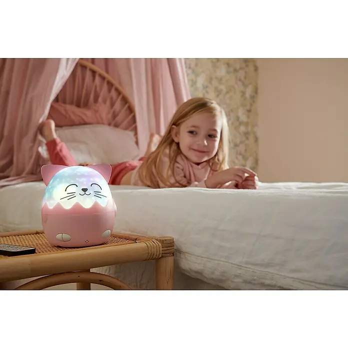 Lámpara de sobremesa LED Mía Gato - Rosa, RGBW - Una linda luz nocturna con forma de gato con proyección de estrellas en una mesita de noche, al lado de una niña acostada en la cama.