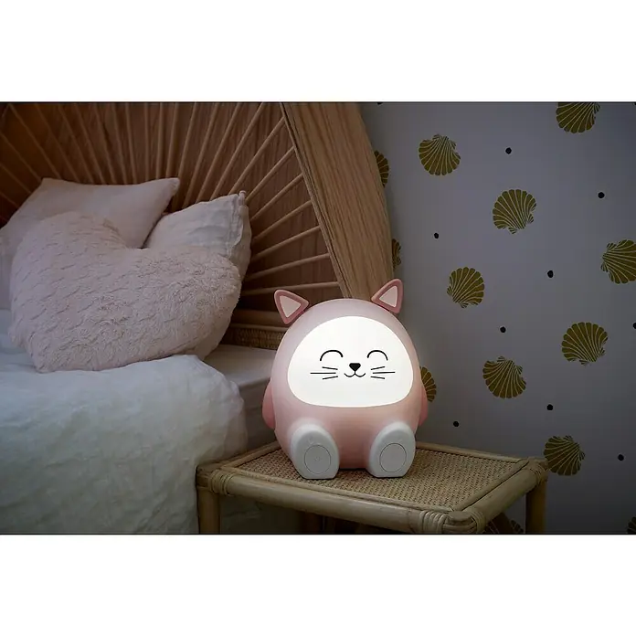 Altavoz Bluetooth Gato Mia - Rosa, 15 W - Luz de noche rosa con forma de gato en una mesita de noche junto a una cama. La luz tiene una cara amable y una luz suave.