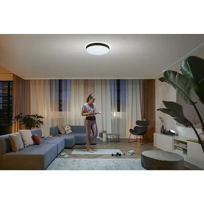 Philips Hue LED-Deckenleuchte rund White Ambience Enrave XL - 48 W, Ø x H: 55,1 x 8,4 cm, Schwarz, Kaltweiß - Eine moderne, flache Deckenleuchte mit Rahmen in einem hell erleuchteten Wohnraum, wo eine Frau Yoga macht. Elegante Beleuchtung für Zuhause.