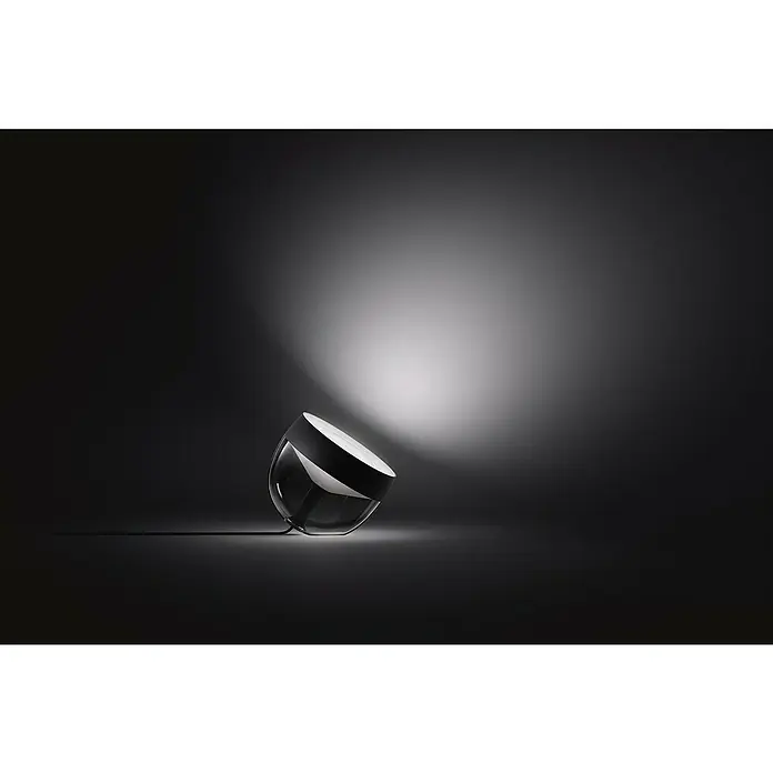 Philips Hue LED-Tischleuchte Iris - Schwarz/Transparent, Höhe: 19,4 cm - Nahaufnahme einer modernen, geneigten Schreibtischlampe mit einem schlanken, schwarzen Lampenschirm und einem klaren Glaskörper, die Licht ausstrahlt.