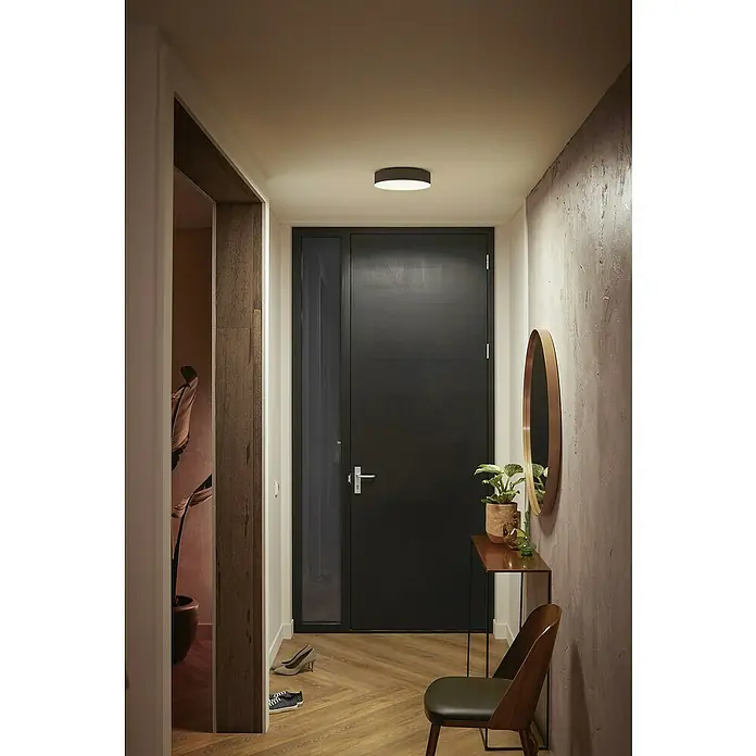 Philips Hue LED-Deckenleuchte rund White Ambience Enrave S - 10 W, Ø x H: 26,1 x 6,6 cm, Schwarz, Kaltweiß - Das Bild zeigt eine moderne Deckenleuchte in einem Flur mit dunkler Tür, Spiegel und minimalistischen Möbeln. Die Leuchte sorgt für angenehmes Licht.