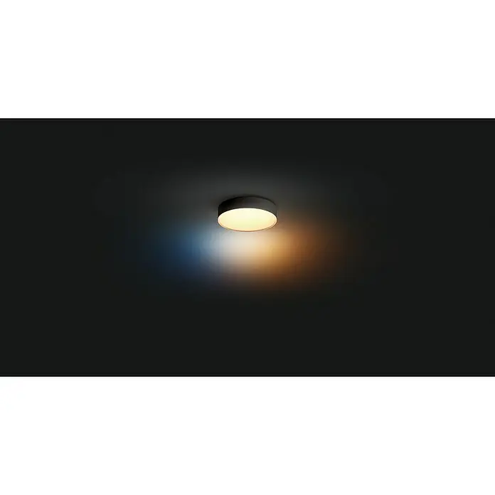 Philips Hue LED-Deckenleuchte rund White Ambience Enrave S - 10 W, Ø x H: 26,1 x 6,6 cm, Schwarz, Kaltweiß - Deckenleuchte in einem dunklen Raum, die ein warmes, gelbes Licht ausstrahlt, das einen angenehmen Kontrast zur Dunkelheit bildet.