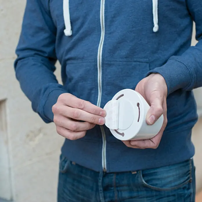 LED-Bewegungsmelder Vigilamp - Weiß, Außen - Eine Person befestigt ein Klebepad an einem runden, weißen Produkt. Das Produkt ist zylinderförmig und weist Schlitze auf.