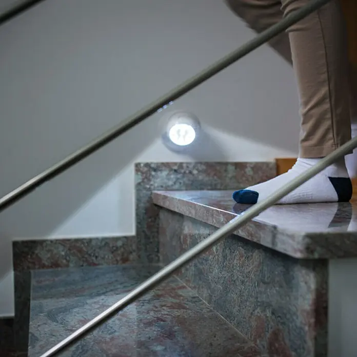LED-Bewegungsmelder Vigilamp - Weiß, Außen - Person, die eine Marmortreppe mit einem Handlauf aus Edelstahl hinaufsteigt, beleuchtet durch eine runde Wandleuchte.