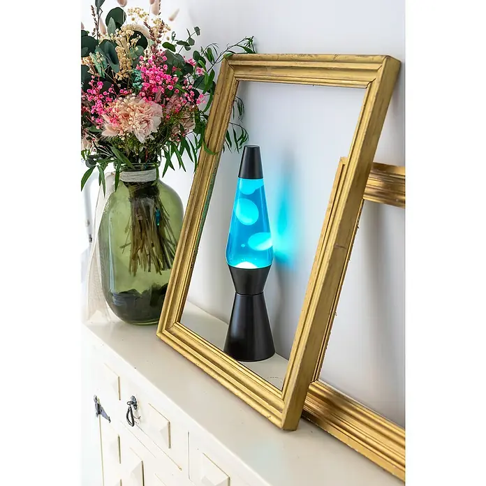 Lámpara de sobremesa decorativa de lava Bullet - 25 W, L x An x Al: 11,5 x 11,5 x 40 cm, Negro - Primer plano de una lámpara de lava azul con base negra, enmarcada por un marco dorado y flores sobre un mueble blanco.