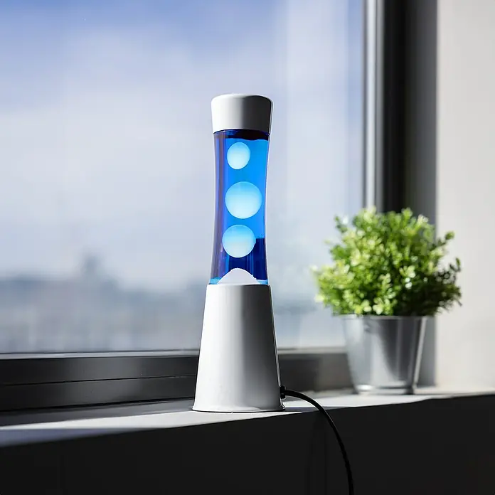 Lámpara de sobremesa decorativa lava mini - 25 W, L x An x Al: 9 x 9 x 30 cm, Blanco, Azul - Una lámpara de lava azul con base blanca y tres burbujas de cera flotantes se sienta en un alféizar de ventana. Una iluminación decorativa para un ambiente acogedor.