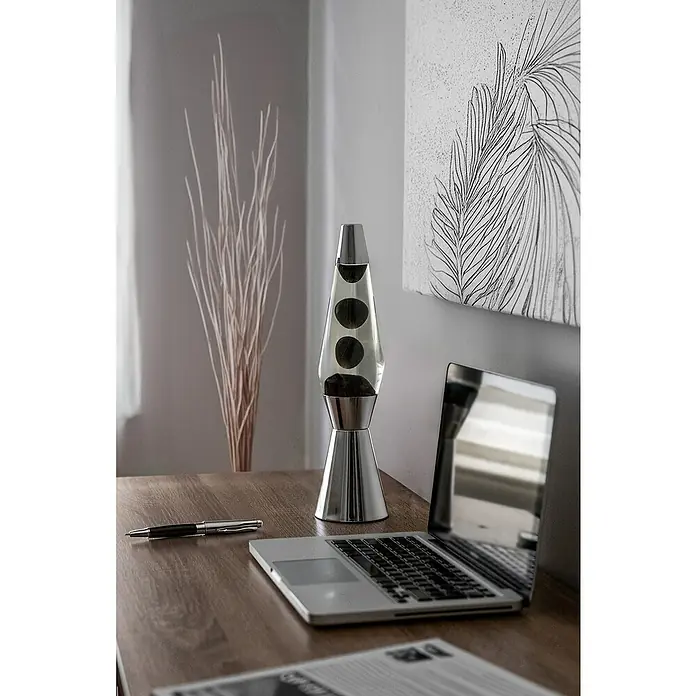 Lámpara de sobremesa decorativa de lava Bullet - 25 W, L x An x Al: 11,5 x 11,5 x 40 cm, Plata - Lámpara de lava plateada con burbujas de cera negra en un escritorio, junto a una computadora portátil y un bolígrafo. Elementos decorativos al fondo.