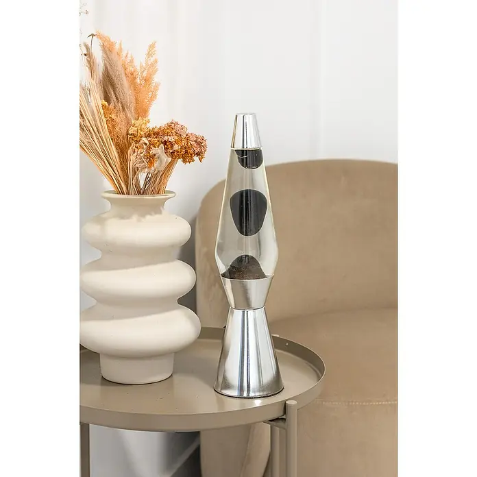 Lámpara de sobremesa decorativa de lava Bullet - 25 W, L x An x Al: 11,5 x 11,5 x 40 cm, Plata - Primer plano de una lámpara de lava con cera negra en una mesa auxiliar, junto a un florero, creando un ambiente cálido y nostálgico.