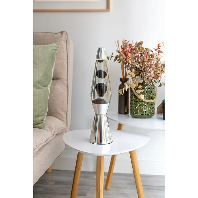 Lámpara de sobremesa decorativa de lava Bullet - 25 W, L x An x Al: 11,5 x 11,5 x 40 cm, Plata - Una lámpara de lava con base plateada y cilindro transparente que contiene burbujas de cera negra sobre una mesa blanca. Jarrón decorativo al fondo.